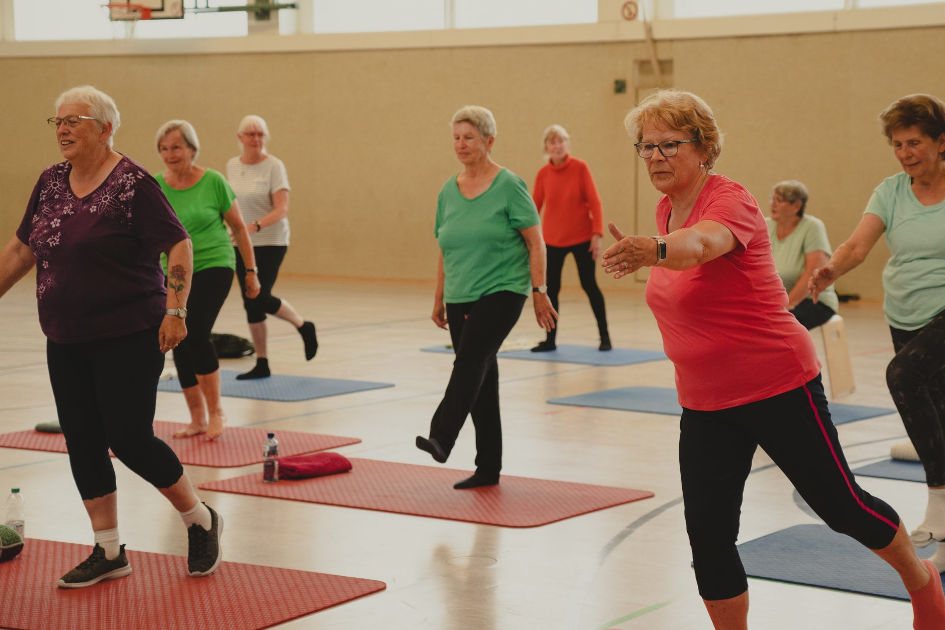 Eine Gruppe älterer Menschen nimmt an einem Fitnesskurs teil. Sie stehen auf Yogamatten in einer Turnhalle und führen Balanceübungen durch. Die Teilnehmer tragen legere Sportkleidung, und der Raum ist gut beleuchtet mit großen Fenstern.