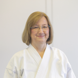 Eine Person trägt eine weiße Kampfsportuniform, auch bekannt als Gi. Das Gesicht der weiblichen Person lächelt. Der Hintergrund ist schlicht und hell.