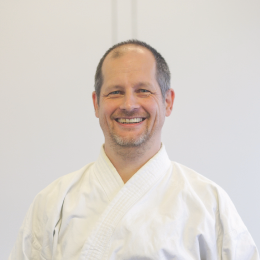 Eine Person trägt eine weiße Kampfsportuniform, auch bekannt als Gi. Der Hintergrund ist schlicht und hell. Die männliche Person lächelt