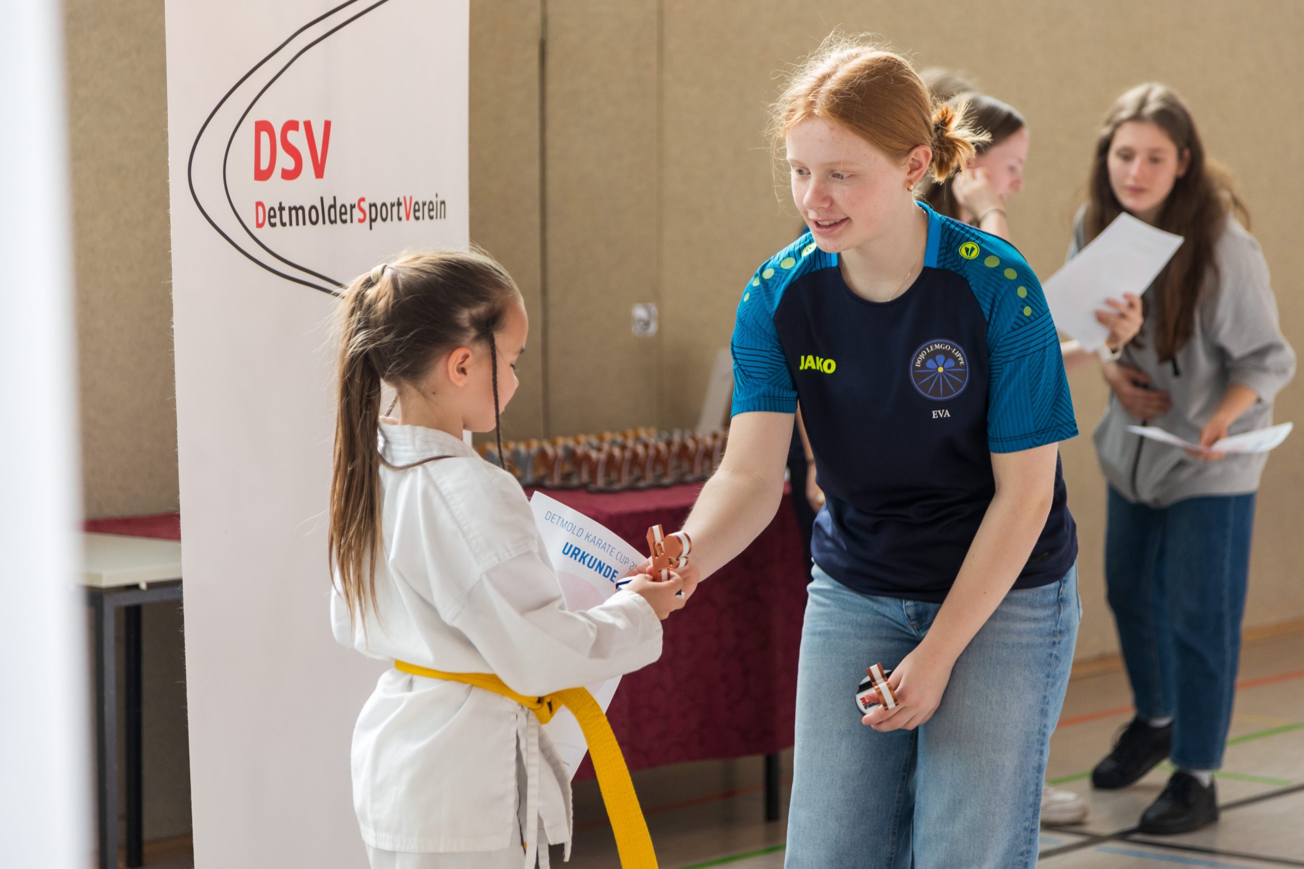 Das Bild zeigt ein junges Mädchen in einer weißen Kampfsportuniform mit einem gelben Gürtel, das von einem Erwachsenen in einem blau-grünen Sportshirt eine Urkunde und eine bronzene Trophäe erhält. Im Hintergrund sind zwei weitere Personen zu sehen, die Urkunden halten. Außerdem gibt es ein Banner mit der Aufschrift "DSV Detmolder Sport Verein