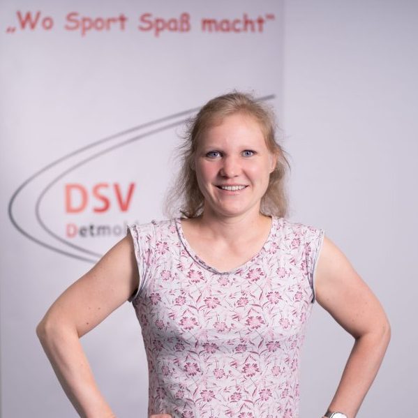 Eine Person steht mit den Händen in die Hüften gestützt vor einem Hintergrund mit dem Schriftzug ‚Wo Sport Spaß macht‘ und ‚DSV Detmold‘. Die Person trägt ein ärmelloses Oberteil mit Blumenmuster.