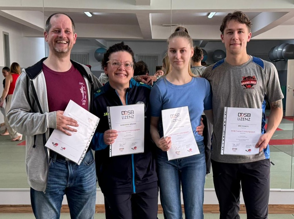 Vier neue Karate-Trainer stehen nebeneinander in einem Sportraum und halten jeweils ein Zertifikat mit der Aufschrift ‚DOSB LIZENZ‘. Die Personen tragen sportliche Kleidung und stehen eng beieinander