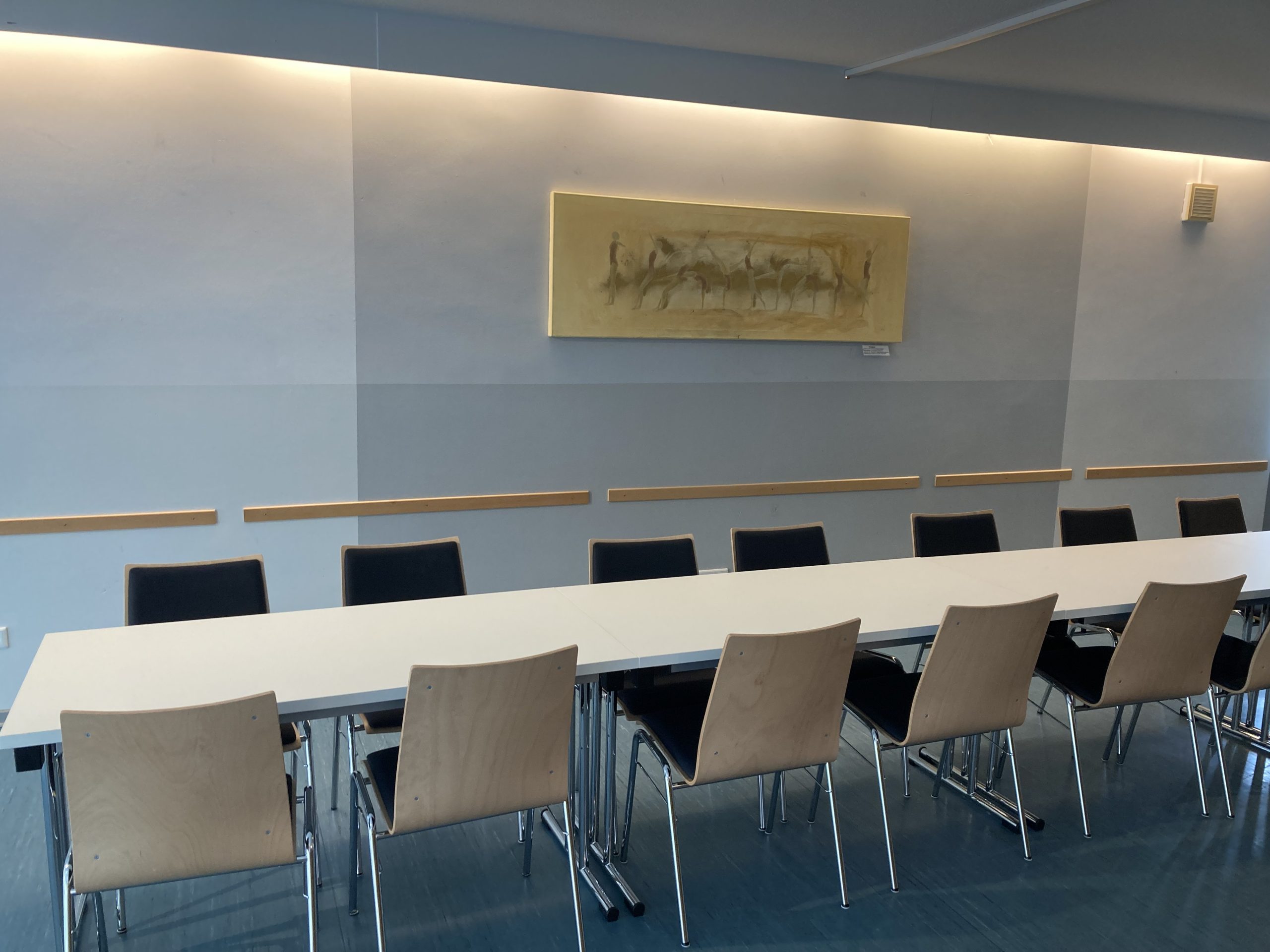 Ein Konferenzraum mit einem langen weißen Tisch und mehreren Stühlen, die darum herum angeordnet sind. Auf einer Seite des Tisches stehen sechs schwarz bezogene Stühle und auf der anderen Seite fünf Stühle mit holzfarbener Rückenlehne. Der Raum hat ein minimalistisches Design mit hellen Wänden und einem Kunstwerk, das über dem Tisch an der Wand hängt. Die Beleuchtung im Raum ist sanft und kommt von Leuchten entlang der oberen Kante der Wände.