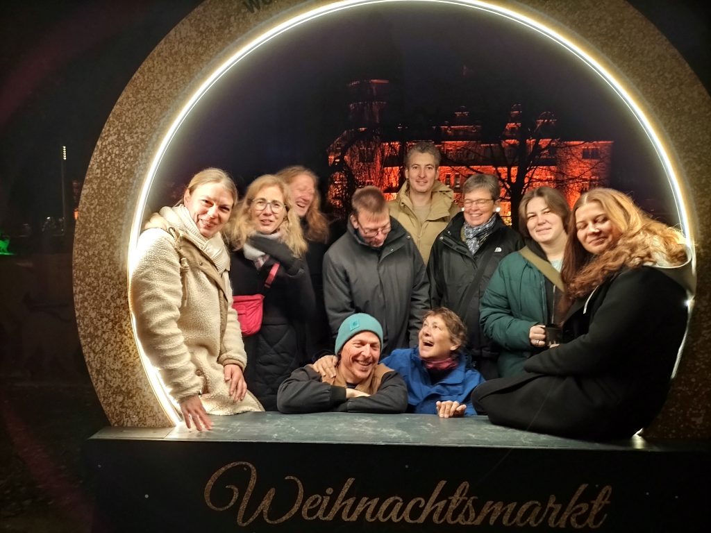 Eine Gruppe von Menschen steht und sitzt in einer großen beleuchteten Ringinstallation auf einem Weihnachtsmarkt. Im Hintergrund ist ein beleuchtetes Gebäude zu sehen.