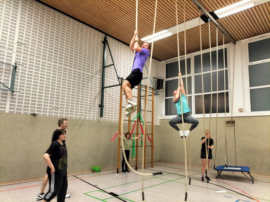 Mehrere Personen trainieren in einer Sporthalle an Kletterseilen. Zwei Teilnehmende klettern aktiv an den Seilen hoch, während andere am Boden warten oder zusehen.