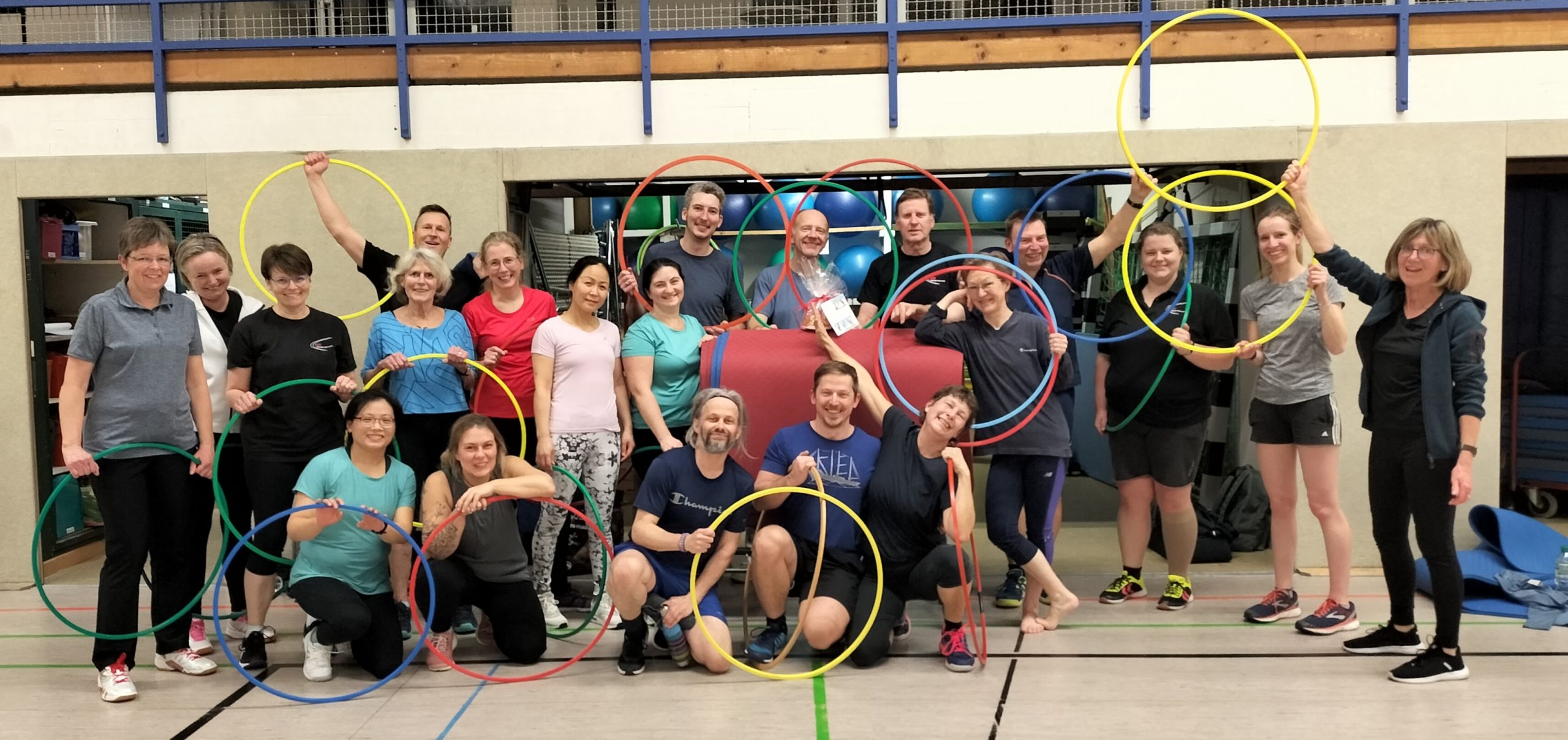 Eine große Gruppe von Menschen steht in einer Sporthalle und hält bunte Hula-Hoop-Reifen in verschiedenen Positionen hoch. Matten und Sportgeräte sind im Hintergrund sichtbar.