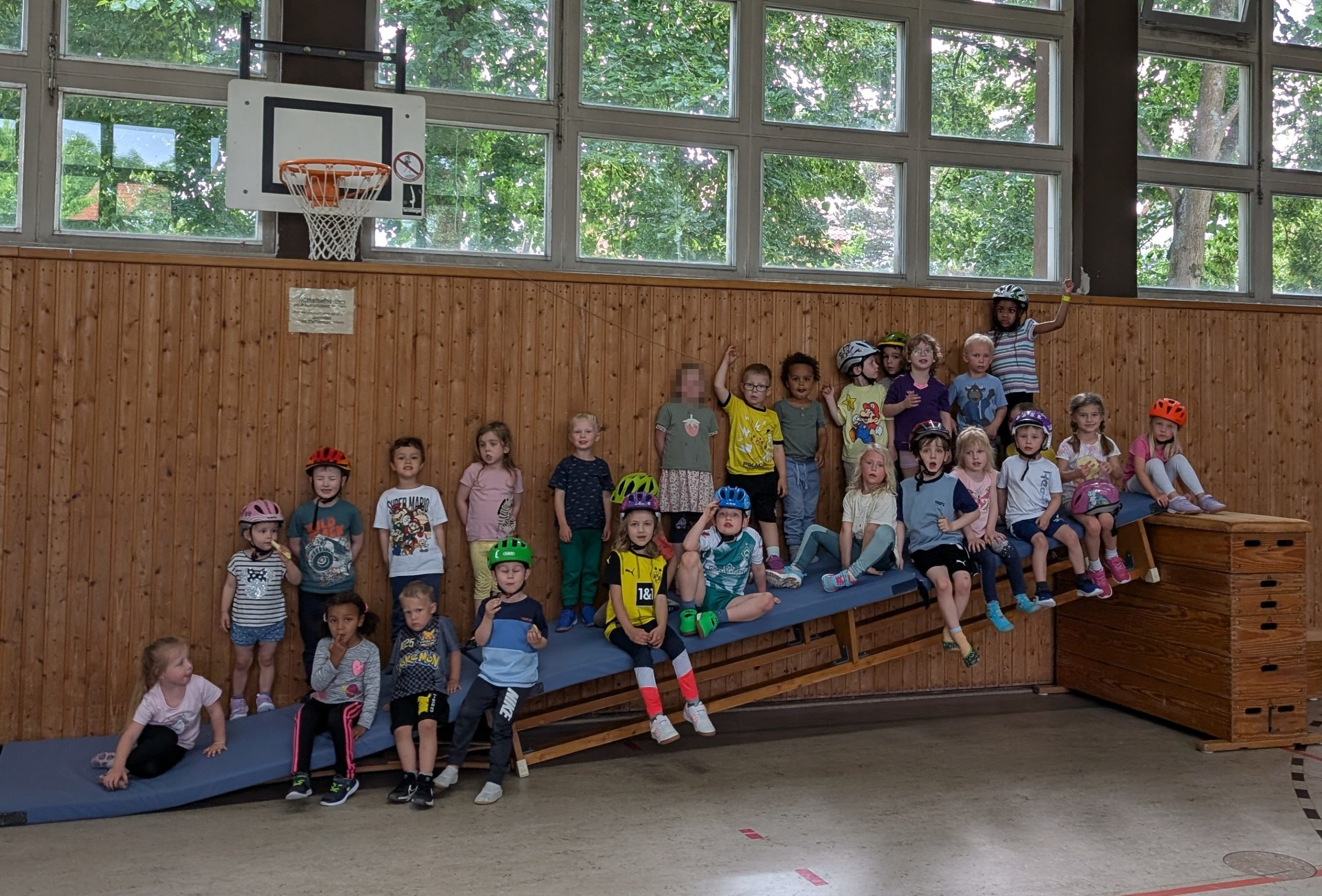 Eine Gruppe von Kindern sitzt und steht in einer Turnhalle auf und neben einer Holzbank, einige tragen bunte Fahrradhelme. Im Hintergrund ist eine Holzvertäfelung und darüber Fenster zu sehen.