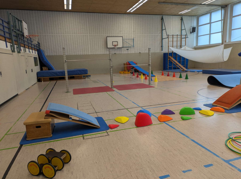 Ein umfangreicher Bewegungsparcours in einer Sporthalle mit bunten Hütchen, Balanciersteinen, einer Rutsche und weiteren Turngeräten. Auf dem Boden sind Turnmatten und Markierungen ausgelegt.