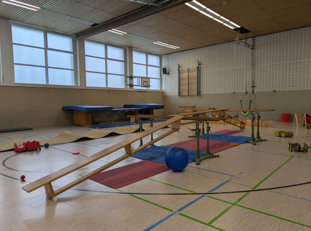 Ein Geräteparcours in einer Sporthalle. Mehrere Holzbänke sind schräg zu Balancierstationen aufgebaut, dazu Matten, Bälle und verschiedene Kleingeräte. Große Fenster lassen Tageslicht in die Halle.