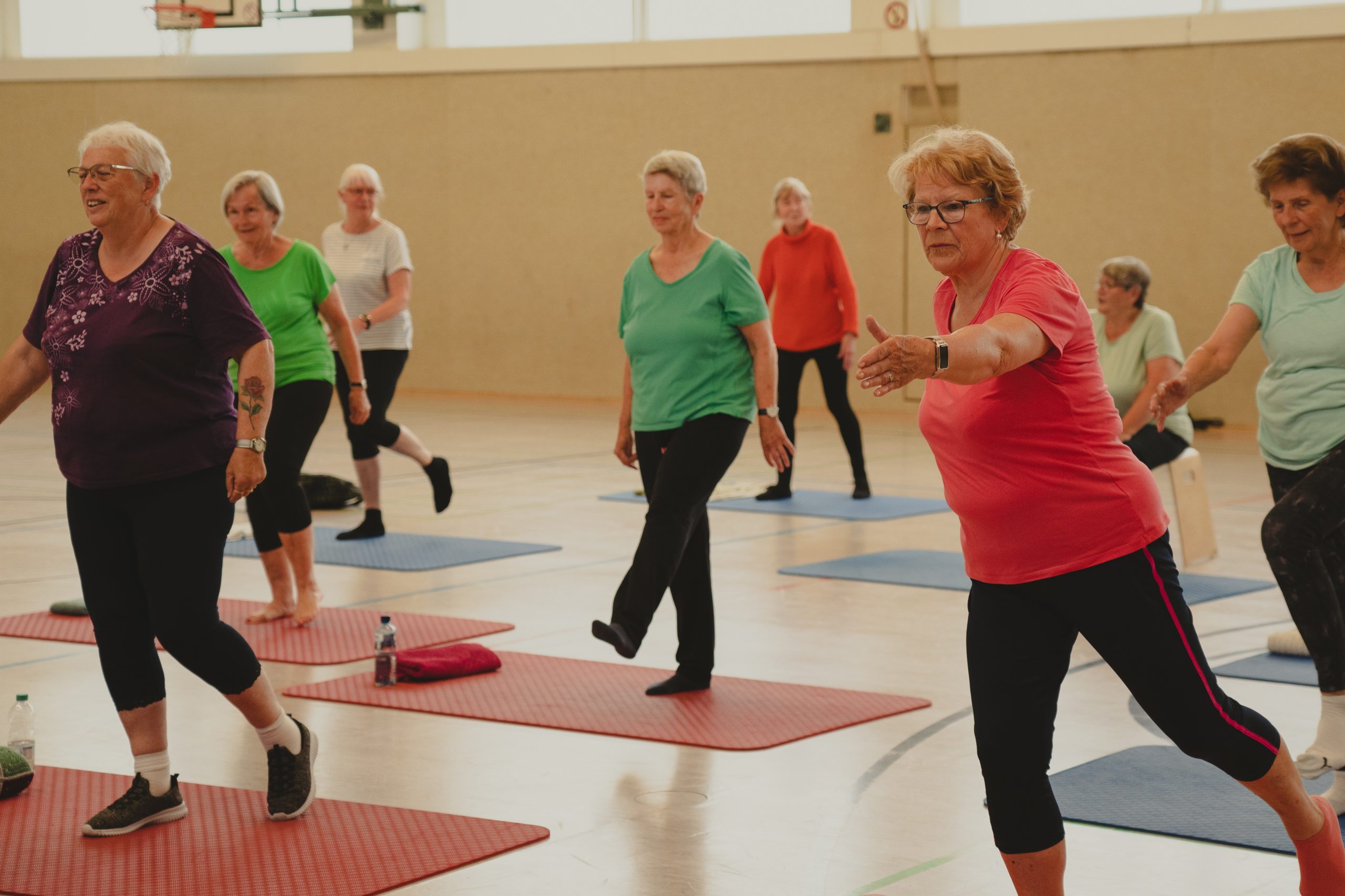 Eine Gruppe von Menschen macht gemeinsam Gymnastik in einer Sporthalle. Sie stehen auf Matten und führen Gleichgewichts- und Dehnübungen aus.