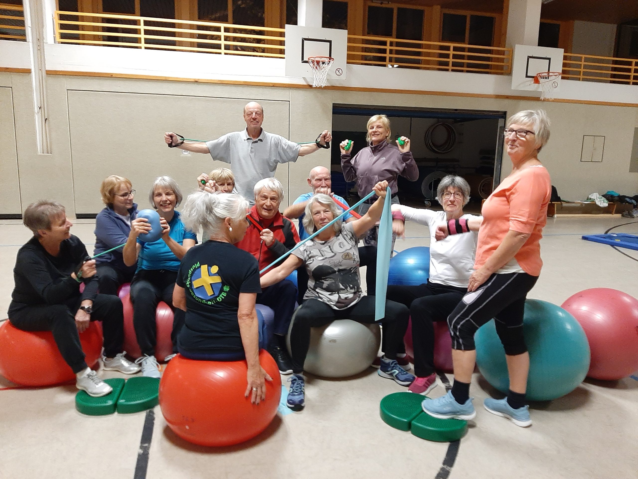 Gruppe von Personen in einer Sporthalle, die gemeinsam Gymnastiktraining macht. Mehrere Personen sitzen auf großen Gymnastikbällen und nutzen Therabänder sowie kleine Hanteln. Eine weitere Person steht seitlich und leitet die Übung an. Im Hintergrund sind Basketballkörbe und Sportgeräte zu sehen.