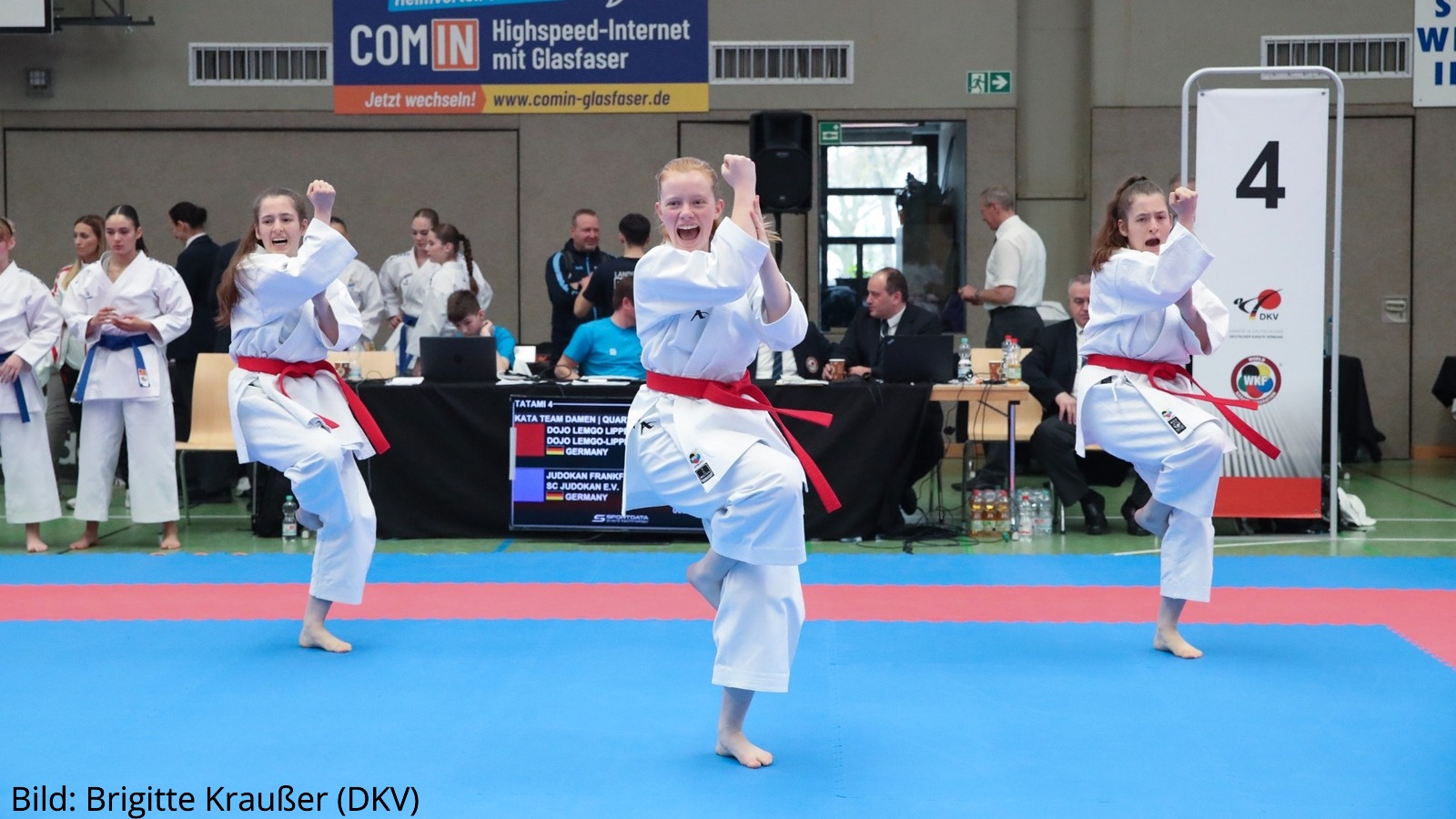 Drei Karateka in weißen Karateanzügen mit roten Gürteln führen synchron eine Kata auf einer blauen Wettkampffläche in einer Sporthalle aus. Im Hintergrund sind Kampfrichtertische, Zuschauer, eine digitale Anzeigetafel sowie Werbebanner zu sehen; rechts steht eine große Tafel mit der Startnummer „4“.